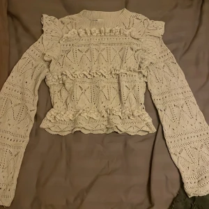 Beige stickad blus med volanger - Superfin beige stickad blus med långärmade ärmar och volangdetaljer på axlar, bröst och midja. Blusen har ett snyggt hålmönster och ribbad halsringning. Perfekt för dig som gillar romantisk och unik stil!