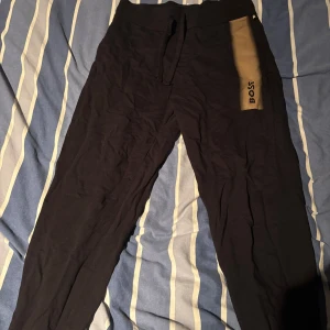Svarta mjukisbyxor från BOSS - Svarta mjukisbyxor från BOSS med snörning i midjan och en beige panel med BOSS-logga på sidan. Byxorna har en slim passform och är tillverkade i mjukt bomullsmaterial, perfekta för chill eller träning. Har ALDRIG använt byxorna och de är i nyskick, använde enbart zip hoodie som medföljde i tracksuiten. 