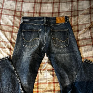 Mörkblå jeans från Jack & Jones - Snygga mörkblå jeans från Jack & Jones med klassisk femficksdesign och slitningar för en cool look. Jeansen har raka ben och normal passform, med detaljerade sömmar på bakfickorna och läderpatch i midjan. Perfekta för dig som gillar stilrena jeans.