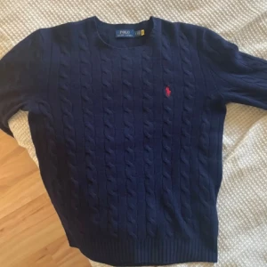 Marinblå kabelstickad tröja Polo Ralph Lauren - Snygg marinblå kabelstickad tröja från Polo Ralph Lauren i mjuk ull och kashmir. Tröjan har rund hals, lång ärm och den klassiska röda Polo-loggan broderad på bröstet. Perfekt för dig som gillar stilrena och tidlösa plagg.
