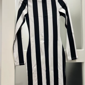 Svartvit randig klänning  - Säljer en svartvit klänning med breda vertikala ränder. Modellen är kort, har lång ärm och rund hals. Materialet är mjukt och stretchigt, troligen bomull med inslag av syntet. Dragkedja i ryggen för enkel påklädning. Slim fit som framhäver figuren.