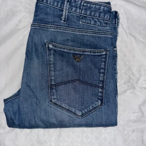 Emporio Armani blå jeans - Snygga blå jeans från Emporio Armani med klassisk femficksdesign och ikonisk logga på bakfickan. Jeansen har normal passform och är tillverkade i mjukt denim-material med knappstängning framtill. Perfekta för dig som gillar stilrena och exklusiva jeans.