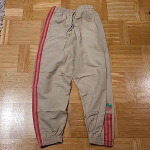 Beige Adidas byxor med röda ränder - Snygga beige Adidas byxor med klassiska röda ränder längs sidorna och broderad Adidas-logga i rött och grönt vid benet. Byxorna har resår och snörning i midjan samt resår vid bensluten. Perfekta för chill eller träning.