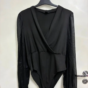 Svart omlott bodysuit med meshärmar - Säljer en svart omlott bodysuit med transparenta meshärmar och v-ringad front. Toppen har ribbad struktur på kroppen och långa, puffiga ärmar i tunn mesh som ger en cool kontrast. Perfekt för dig som gillar stilrena och trendiga plagg med lite edge.