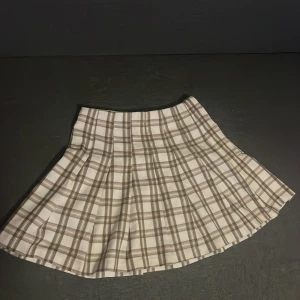 Beige rutig plisserad kortkjol - Snygg beige och vit rutig plisserad kortkjol med klassisk skolstil. Kjolen har en A-linjeformad siluett och är tillverkad i ett mjukt bomullsmaterial som känns skönt mot huden. Perfekt för dig som gillar preppy vibes och vill sticka ut med ett trendigt mönster.