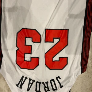 Chicago Bulls Jordan 23 fotbollströja - Vit fotbollströja med svarta och röda detaljer, Chicago och nummer 23 i rött framtill. Sidorna har svarta och röda ränder. Materialet är mesh för bra andningsförmåga. Silverfärgad logga på framsidan och klassisk Jordan-stil.