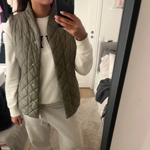 Olivgrön quiltad väst från Lindex - Quiltad olivgrön väst från Lindex med snyggt rutmönster och tryckknappar framtill. Västen har en rak passform och är tillverkad i polyester, perfekt att slänga över hoodien för en chill look. Fickor på sidorna för extra funktion.