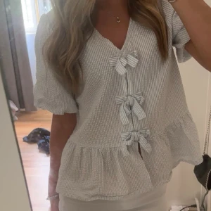 Vit blus med rosetter och volang - Supersöt vit blus med tunna blå ränder, v-ringning och tre knytrosetter framtill. Blusen har puffiga korta ärmar och volang nertill för en romantisk vibe. Perfekt till jeans eller kjol för en fräsch look.