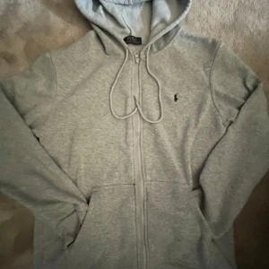 Grå hoodie från Polo Ralph Lauren - Säljer en stilren grå hoodie från Polo Ralph Lauren med dragkedja och justerbar huva. Den har en broderad logga på bröstet och är tillverkad i mjukt material.