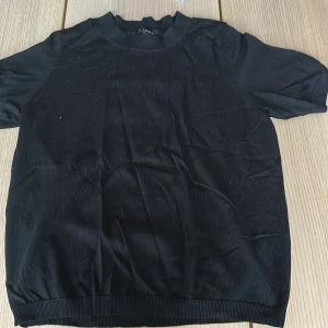 Svart kortärmad tröja från Lindex XS - En svart, enkel och stilren kortärmad tröja från Lindex i storlek XS. Tröjan har ribbad halsringning och nederkant, och är gjord i ett mjukt material som känns skönt mot huden. Perfekt till jeans eller kjol för en clean look.