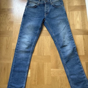 Blå jeans med raka ben från Nudie - Snygga blå jeans från Nudie med klassisk femficksdesign och raka ben. Jeansen har en mörkblå tvätt med lätt slitning och Nudies ikoniska broderi på bakfickorna. Passar dig som gillar en enkel och tidlös look.