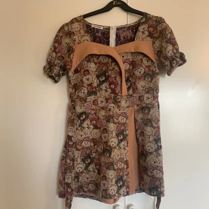 Kort klänning med nallemönster från Nuy Shop - Unik kort klänning från Nuy Shop med ett färgglatt nallemönster i beige, brun och rosa toner. Klänningen har puffiga korta ärmar, ett bälte i midjan och  champagnefärgade detaljer framtill. Materialet är tjockt och vävt, perfekt för dig som gillar statement-plagg.