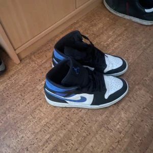 Nike Air Jordan 1 blå/svart sneakers - Nike Air Jordan 1 sneakers i blått, svart och vitt med klassisk high-top siluett. Skorna har snörning, rund tå och platt sula. Ikonisk Jumpman-logga på plösen och Nike swoosh på sidan. Perfekt för dig som vill ha en streetstyle vibe med retro känsla.