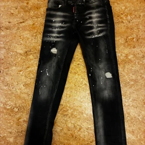 Svarta skinny jeans med slitningar - Säljer ett par svarta skinny jeans från Dsquared2 med coola slitningar, färgstänk och distressed detaljer på framsidan. Jeansen har en smal passform, klassisk femficksdesign och är tillverkade i stretchig bomull för skön komfort.