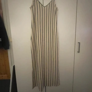 Randig långklänning från Gina Tricot - Långklänning från Gina Tricot i beige med svarta vertikala ränder. Klänningen har smala axelband, v-ringning och är ärmlös. Materialet är ribbat och känns mjukt mot huden. Perfekt för en trendig och avslappnad look.