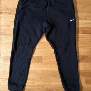 Svarta mjukisbyxor från Nike - Svarta mjukisbyxor från Nike med klassisk logga på vänster ben. Byxorna har en slim passform med muddar vid bensluten och elastisk midja. Perfekta för chill eller träning, tillverkade i mjukt bomullsmaterial för extra komfort.