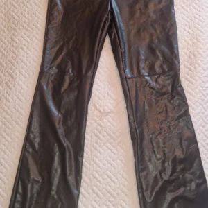 Svarta bootcut byxor i skinnimitation - Snygga svarta byxor i skinnimitation med bootcut passform. Byxorna har en varm och skön insida och har en slät yta och markerad söm vid knäna som ger en cool detalj. Perfekta för dig som vill ha en edgy look och gillar att sticka ut.