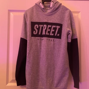 Grå hoodie med svart tryck STREET. - Snygg grå hoodie med svart tryck där det står STREET. och texten STAY TRUE under. Tröjan har svarta ärmar, huva med vita snören och är tillverkad i mjuk bomullsblandning. Perfekt för en avslappnad streetstyle-look.