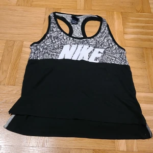 Nike svart och vit linnetopp XS - Nike linnetopp i storlek XS med svart nedre del och vit/svart grafiskt mönster upptill. Stort Nike-tryck framtill. Sportig racerback och ärmlös design, perfekt för träning. Materialet är mjukt och lätt polyester som andas.