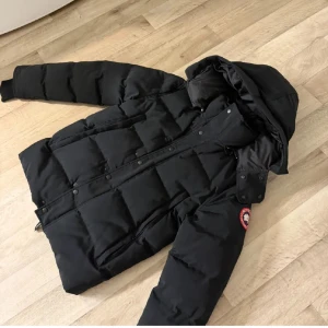 Svart dunjacka från Canada Goose M - Säljer en svart dunjacka från Canada Goose i storlek M. Jackan har huva, quiltad design och klassisk logga på ärmen. Perfekt för kalla dagar med sin tjocka och värmande fyllning. Stängs med dragkedja och tryckknappar framtill. Grym stil för vintern!