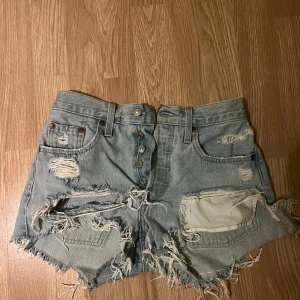 Ljusa slitna jeansshorts med knappar - Riktigt snygga ljusblå jeansshorts med flera slitningar och fransiga kanter. Knappar framtill istället för dragkedja och klassiska fickor både fram och bak. Perfekta för en cool och avslappnad stil i sommar.