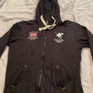 Svart hoodie från Vinson Polo Club - Svart hoodie från Vinson Polo Club med broderad logga och text på bröstet. Tröjan har vit snörning i huvan, dragkedja framtill och två fickor med vita detaljer. Materialet är mjukt och känns skönt mot huden. Perfekt för chill dagar och streetwear.
