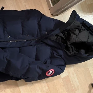 Mörkblå dunjacka från Canada Goose - Säljer en mörkblå dunjacka från Canada Goose med huva och klassisk logga på ärmen. Jackan har dragkedja framtill och är fodrad med dun för extra värme. Perfekt för kalla dagar och har en clean, stilren look som funkar till det mesta.