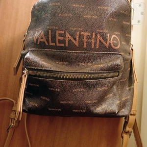 Brun ryggsäck från Valentino - Snygg brun ryggsäck från Valentino med logomönster över hela väskan och stora bokstäver framtill. Väskan har dragkedja, justerbara axelremmar i beige och flera fack både utvändigt och invändigt. Perfekt för dig som vill ha en trendig och praktisk väska.