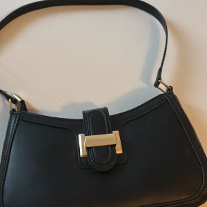 Svart axelväska med gulddetaljer - Stilren svart axelväska i skinnimitation med guldfärgat spänne framtill. Väskan har en smal axelrem och stängs med dragkedja. Perfekt storlek för det viktigaste och ger en snygg touch till din outfit.