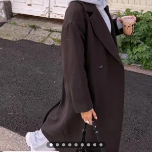 Mörkbrun lång kappa med knappar slutsåld junkyard - Stilren mörkbrun kappa med klassisk krage och enkel knäppning framtill. Lång modell med rak passform och diskret slits bak. Perfekt för dig som gillar en clean och tidlös look. Helt slutsåld på junkyard. Brukar ha S men denna i XS passar mycket bra. Använd ca 10 gånger därav i super skick