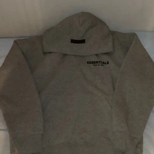 Grå Essentials Fear of God  - Snygg grå hoodie från Essentials Fear of God med stor svart logga på ryggen och mindre tryck på bröstet. Tröjan har huva och är tillverkad i mjuk bomull med fleece på insidan. Perfekt för dig som gillar streetwear och vill ha en clean look.