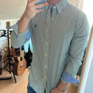 Grön- och vitrandig skjorta från Ralph Lauren - Snygg skjorta med smala gröna och vita ränder från Ralph Lauren. Klassisk button-down krage, broderad logga på bröstet och vita knappar. Ärmsluten har blå insida som syns när de viks upp. Perfekt till jeans för en clean look.