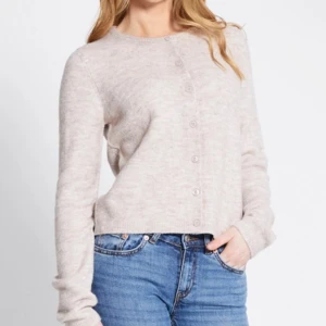 Beige stickad kofta med knappar - Mysig beige stickad kofta med rund hals och knäppning framtill. Koftan har långa ärmar och en mjuk, lite fluffig känsla. Perfekt att slänga på sig när det är lite kyligt ute. Enkel och stilren design som funkar till jeans eller kjol. 