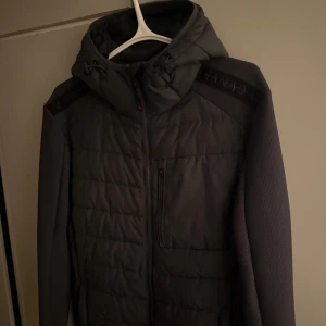 Grå cardigan från Zavetti Canada - Snygg grå cardigan från Zavetti Canada med huva och snörning. Jackan har quiltad framsida, dragkedja och coolt mönstrade axelpartier med logotyp. Perfekt för kalla dagar och har en modern, sportig vibe.