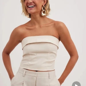 Beige axelbandslös tubtopp med dragkedja - Säljer en stilren beige tubtopp med bred kant upptill och dold dragkedja i ryggen. Toppen är croppad och har en clean, minimalistisk look som passar perfekt till kostymbyxor eller jeans. Gjord i ett slätt tyg med lätt struktur.