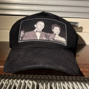 Svart Maggiore keps med mesh och tryck - Säljer en svart keps från Maggiore med mesh på sidorna och baksidan för extra ventilation. Framtill finns ett stort tryck med retrofoto och Maggiore-logga. Justerbar snapback och insida med logotypband. Perfekt för dig som gillar streetwear och unik design.