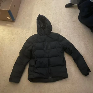 Svart pufferjacka från Everest - Svart pufferjacka från Everest med huva och flera praktiska fickor med dragkedja. Jackan har en stilren look med diskret logga på bröstet och grova sömmar som ger en schysst puffad känsla. Perfekt för kalla dagar och enkel att matcha med din vardagsstil.vid snabb köp kan priset diskuteras modell 175 cm lång 55 kg