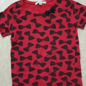 T-shirt - Röd t-shirt från Mywear Young med svarta rosetter som mönster över hela tröjan. Kortärmad modell i mjuk bomull och en söt svart rosett vid halsringningen. Perfekt för dig som gillar lekfull stil och vill sticka ut lite extra.