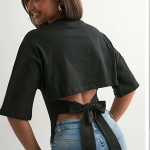 Svart top med knyt i ryggen från Nelly - Säljer en svart topp med halvlånga ärmar och snygg öppen rygg med breda band som knyts i en stor rosett. Toppen har en croppad passform och är tillverkad i ett mjukt, slätt material. 