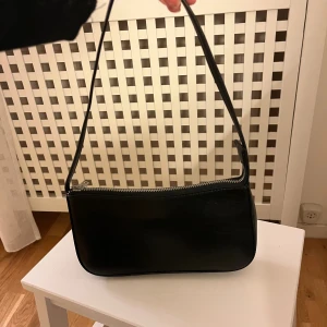 Svart axelväska i skinn med dragkedja - Snygg svart axelväska i skinn med stilren, avlång form och silverfärgad dragkedja upptill. Väskan har ett innerfack med dragkedja och en enkel, minimalistisk design som passar till många olika outfits. Perfekt storlek för det viktigaste.