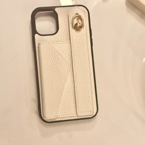 iPhone 11 Plånboksfodral Kortfack - Elegant vitt plånboksfodral till iPhone 11 med strukturmönstrad yta, handrem och guldfärgad detalj. Fodralet har ett praktiskt kortfack med magnetlås. Skicket är mycket gott, utan synligt slitage eller defekter. 