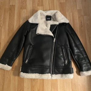 Svart pälsjacka från Pull&Bear - Snygg svart pälsjacka från Pull&Bear med vit fluffig fuskpäls på insidan och krage. Jackan har två dragkedjeförsedda fickor, detaljer med spännen vid midjan och en rejäl asymmetrisk dragkedja framtill. Perfekt för kalla dagar och riktigt stilren look.