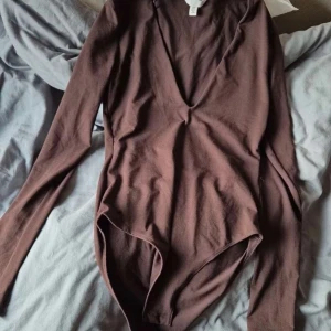 Brun långärmad bodysuit från H&M - Säljer en brun långärmad bodysuit från H&M i mjuk bomull. Modellen har en djup v-ringning och tight passform som sitter snyggt mot kroppen. Perfekt att styla med jeans eller kjol för en clean och enkel look. Säljer pågrund av förstor. Ny utan prislapp! 