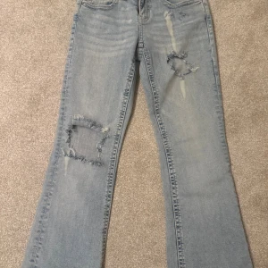 Bootcut ljusblå jeans från Perfect Jeans - Snygga ljusblå bootcut jeans från Perfect Jeans med slitna detaljer och hål på båda benen. Klassisk femficksmodell, låg midja och knappar bak på fickorna. Jeansen har en cool tvättad look och är tillverkade i mjukt denimtyg.