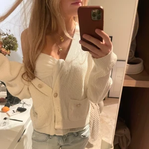 Vit ribbad kofta med fickor - Säljer en vit ribbad kofta med snygga guldfärgade knappar och två praktiska fickor framtill. Koftan har en croppad passform och långa ärmar, perfekt att slänga över en topp. Mjuk stickad känsla och stilren look som funkar till jeans eller kjol.