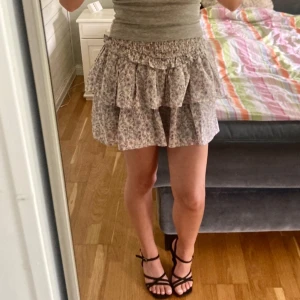 Blommig volangkjol med resårmidja - Supersöt kort kjol med blommigt mönster i ljusa toner. Kjolen har flera volanger och en bred, smockad resår i midjan som gör den bekväm och enkel att bära. Perfekt för dig som gillar romantisk och feminin stil.