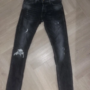 Svarta slim jeans Jack & Jones 29/32 - Svarta slim fit jeans från Jack & Jones, modell Glenn. Jeansen har slitna detaljer och hål på benen för en cool, edgy look. Klassisk femficksdesign och smal passform som sitter snyggt längs benen. Perfekt för dig som gillar streetstyle eller grisch. 29/32 storlek 