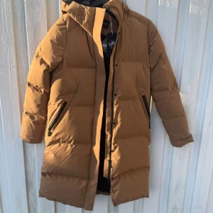 Brun lång dunjacka Sail Racing M - Säljer en brun lång dunjacka från Sail Racing med huva och svarta dragkedjor på fickorna. Jackan har quiltad design, logotyp på ärmen och är perfekt för kalla dagar. Fylld med Allied Feather Down och har en stilren look som funkar till streetwear.