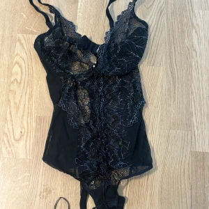 Svart spetsig korsett med snörning - Säljer en svart korsett med genomskinlig spets och snygga detaljer. Den har smala axelband, snörning framtill och hakknäppning bak. Spetsen har ett blommigt mönster och ger en riktigt elegant vibe. Perfekt för dig som vill ha något extra till outfiten.