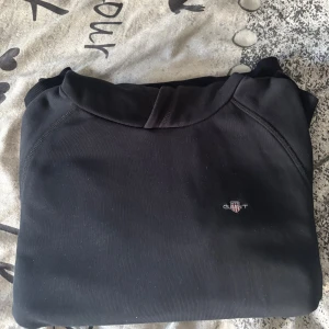 Svart hoodie från GANT med logga - Svart hoodie från GANT med broderad logga på bröstet. Klassisk design med huva och magficka, ribbade muddar och mjukt material. Perfekt för chill dagar och enkel att matcha med jeans eller joggers.